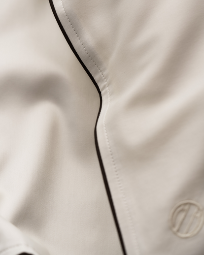 Close-up of a white or off-white fabric garment with a thin black piping trim running vertically, featuring a tonal embroidered logo in the lower right corner.